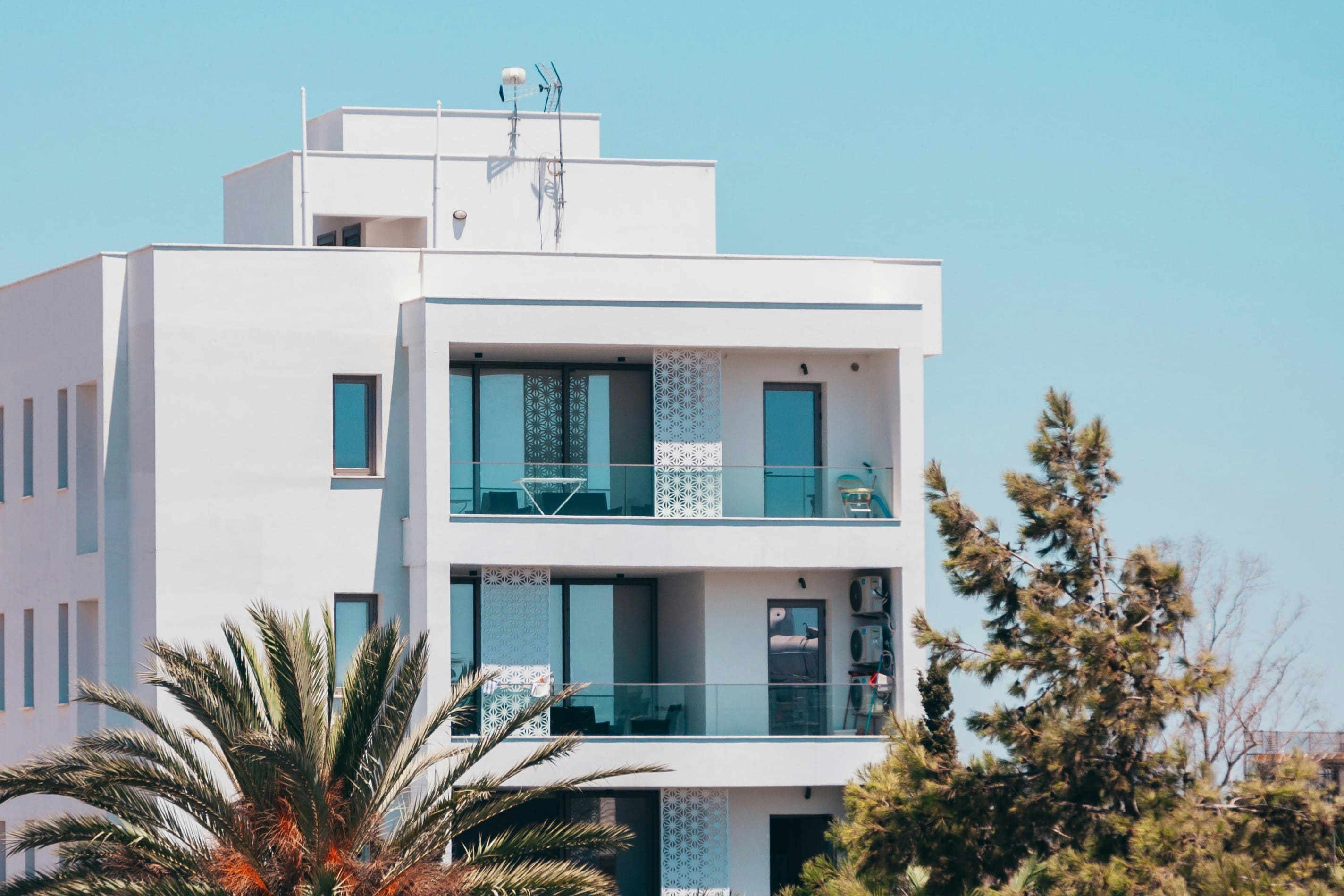 A white, modern apartment building with glass balconies overlooking palm trees and a blue sky.